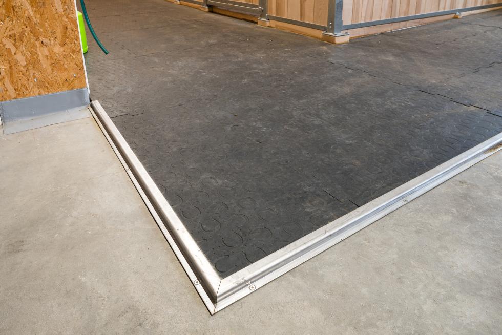Ein Gummimattenweg, endend mit einem z-förmigen Winkel, welcher auf der Gummimatte aufliegt, im rechten Winkel senkrecht der Seite hinunterfolgt und anschliessend wieder durch einen 90° Winkel waagerecht auf dem Boden aufliegt, der Weg macht eine Kurve um eine hölzerne Pferdebox, Draufsicht 